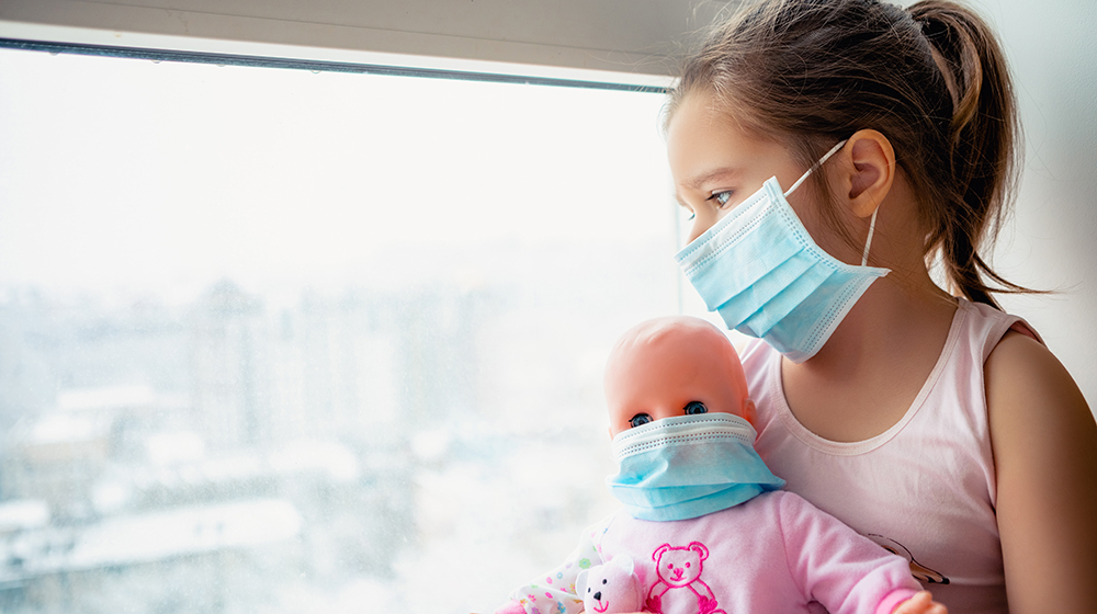 Kleines Mädchen mit OP-Maske sitzt am Fenster mit Puppe im Arm (c) shutterstock : L Julia