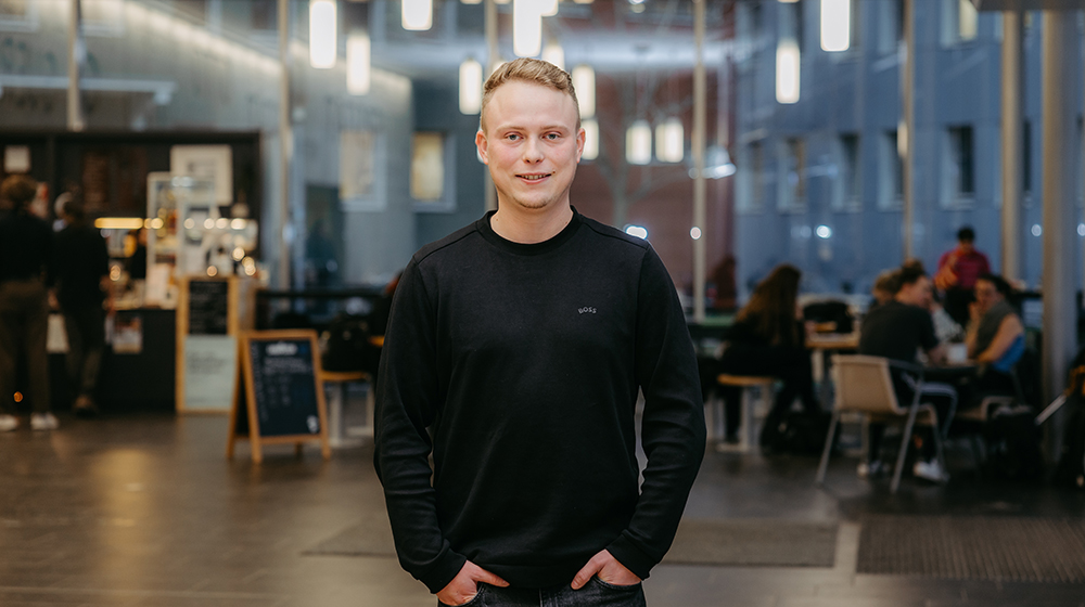 Portrait von Sascha Muth im Foyer von Gebäude 44 mit einem Café und Menschen im Hintergrund (c) Jana Dünnhaupt Uni Magdeburg