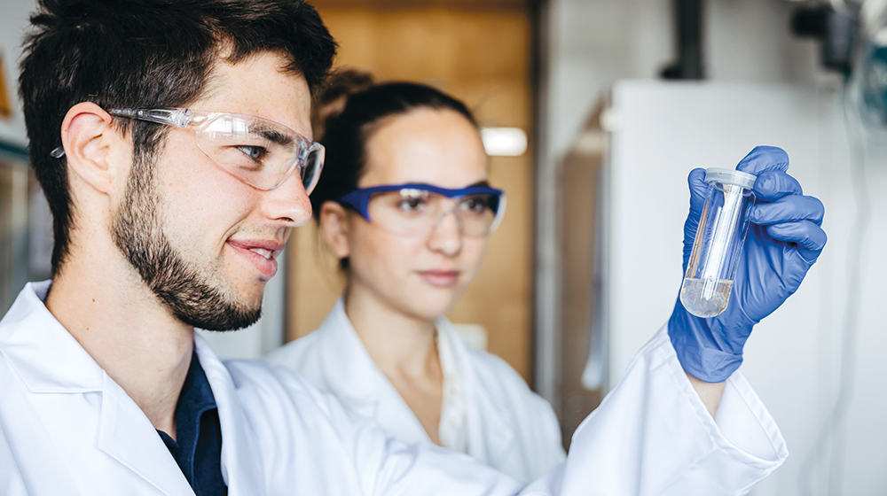 In the lab: M. Sc. Ruben Goldhahn holds a test tube in his hand, next to him stands M. Sc. Ann-Joelle Minor (Photo: Jana Dünnhaupt / University of Magdeburg)